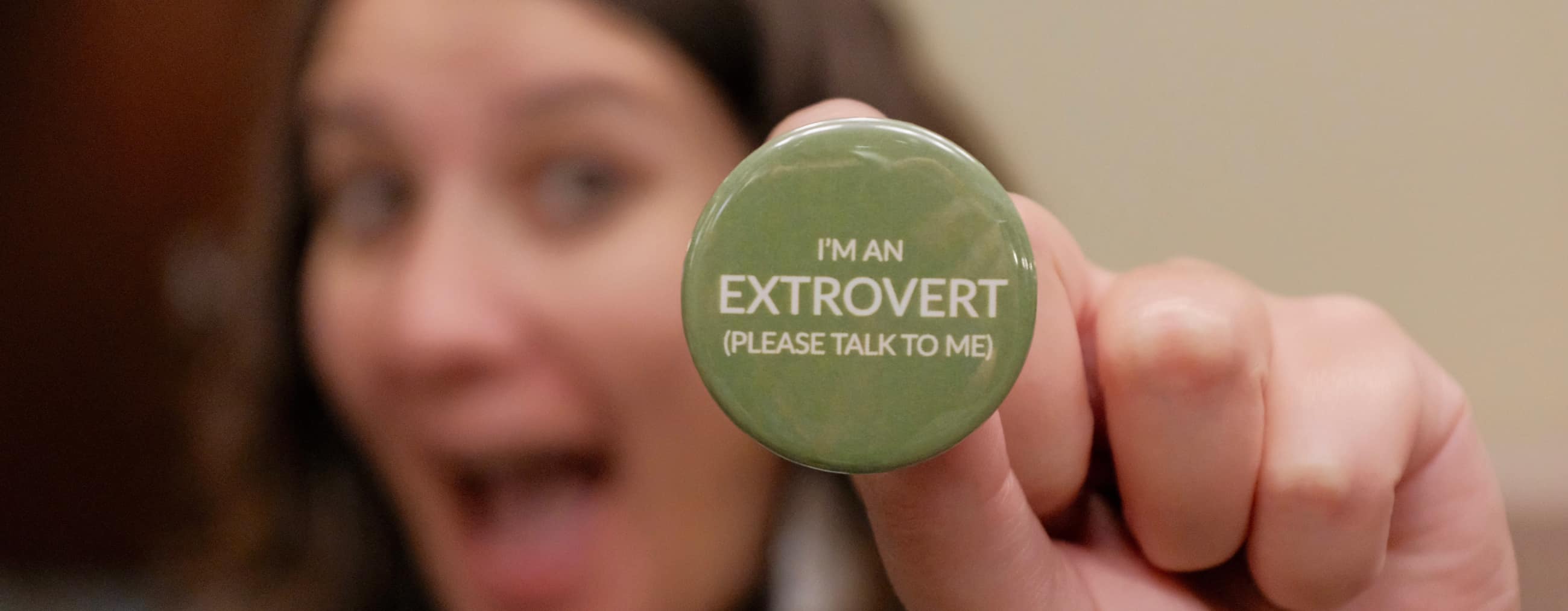 Tenessa holds a green circular pin toward the viewer, reading “I’m an extrovert, please talk to me.” Tenessa is out of focus behind it, with an excited expression.