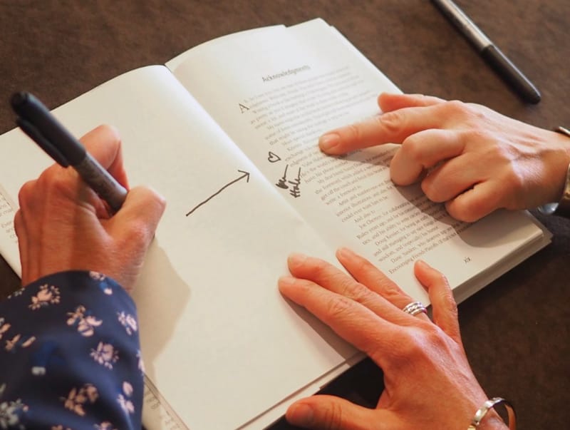 A book held open with one hand, while the other writes notes in the margins with a Sharpie. A second person is pointing at a spot on the page.